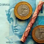 Cédula de R$ 100 onde duas moedas de um real e uma cédula de R$ 10 enrolada formam um símbolo de percentual.