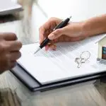 Pessoa assinando contrato de compra com um miniatura de casa sobre os papéis