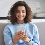 Mulher sentada no sofá segurando um celular. Foto: Adobe Stock
