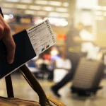 Mão segurando um passaporte com uma passagem aérea em um aeroporto. Foto: Pixabay