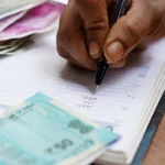 Mão anotando em caderno, com notas em volta. Foto: Pixabay