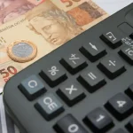 Calculadora com cédulas de Real e moeda de 1 real ao fundo apoiados sobre uma mesa. Foto: Marcello Casal Jr/Agência Brasil