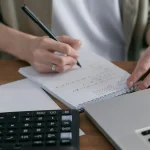Mulher escrevendo em papel com calculadora ao lado. Foto: Pexels
