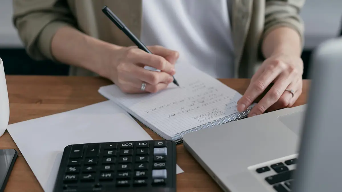 Mulher escrevendo em papel com calculadora ao lado. Foto: Pexels