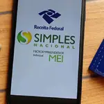Celular apresenta logo da Receita e do Simples Nacional e MEI, juntamente com calculadora e caderno. Ferramentas que auxiliam no cálculo do IR. Foto: Brenda Blossom - Adobe Stock