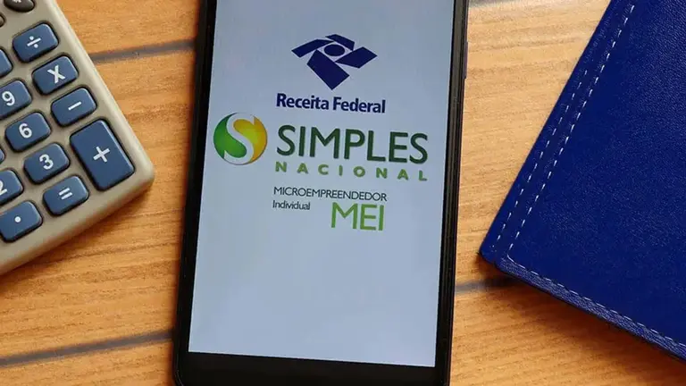 Celular apresenta logo da Receita e do Simples Nacional e MEI, juntamente com calculadora e caderno. Ferramentas que auxiliam no cálculo do IR. Foto: Brenda Blossom - Adobe Stock